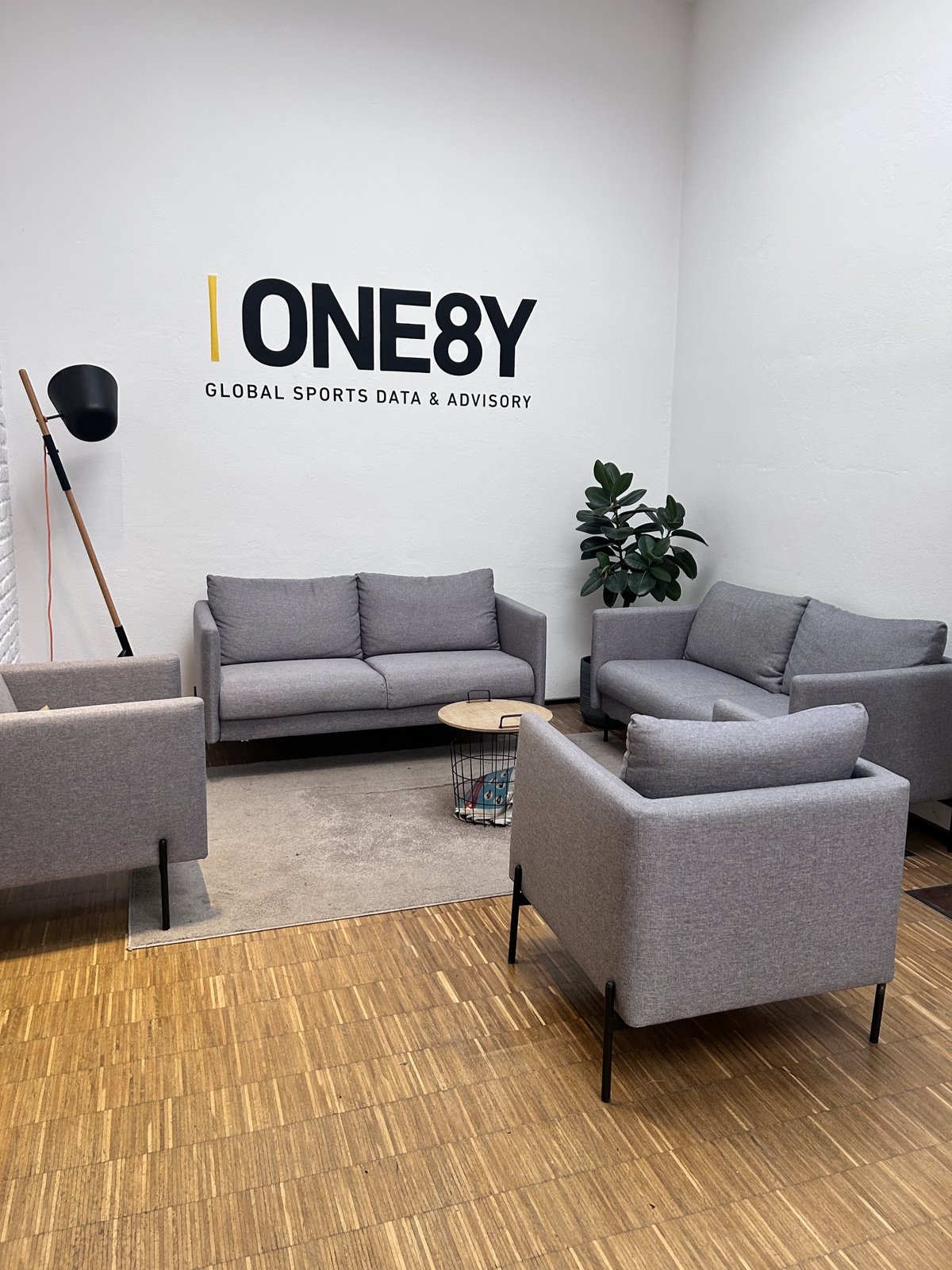 Lounge bei ONE8Y mit grauen Sofas vor dem ONE8Y-Schriftzug Global Sports Data and Advisory